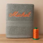 Serviette de bain grise brodée au prénom Michel avec fil orange, création artisanale Sudbroderie à Saint-Cyr-sur-Mer, accompagnée de bobines de fil Madeira.