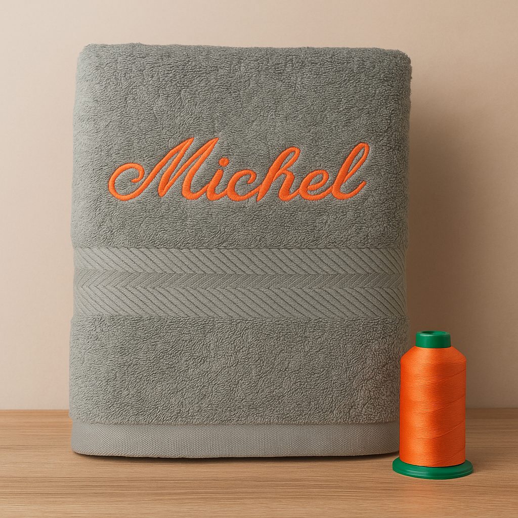 Serviette de bain grise brodée au prénom Michel avec fil orange, création artisanale Sudbroderie à Saint-Cyr-sur-Mer, accompagnée de bobines de fil Madeira.